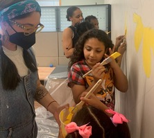 Two women and four children painting yellow paint on a wall.