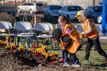 Kids in reflective gear shoveling dirt with toy shovels and dump trucks.