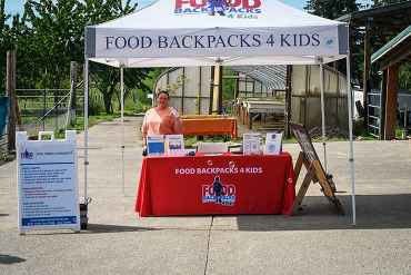 Woman standing at a Food Backpacks 4 Kids outreach table at an event.