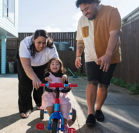 Man and woman helping toddler girl to ride a tricycle.