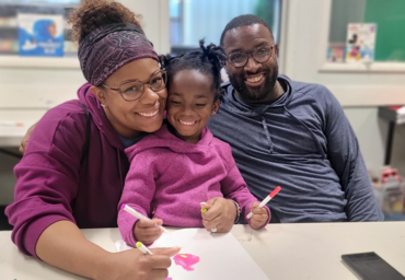Smiling mother and father sitting at a table with their daughter who is coloring with markers.