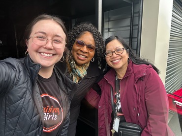 Three smiling women in front of the trunk of an open minivan.