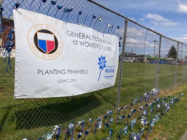 Pinwheel in the lawn in front of a General Federation of Women's Club sign hanging from a chain link fence.