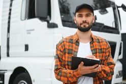 A bearded truck driver in an orange plaid shirt and black cap holds a clipboard and pen in front of a white semi-truck