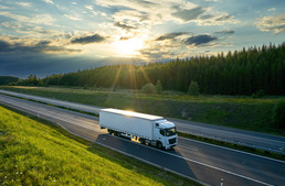 A white semi-truck driving down an empty highway at sunset, surrounded by green fields and a forest in the distance.