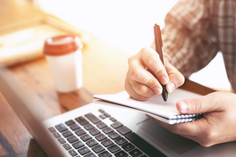 Person writing in a notebook next to a laptop and coffee cup.