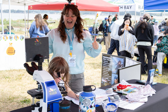 A child looks through a microscope while an adult looks ecstatic behind them. Lots of other people explore booths.