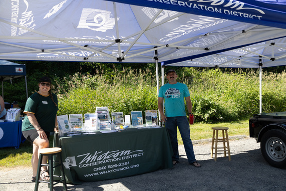 Two staff members stand next to a table and watershed model under Whatcom CD branded tents. Other booths are set up behind them. 