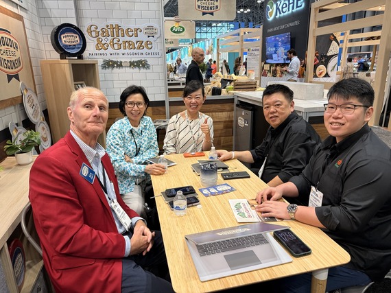 Five people sit around a table with laptops, water bottles, and phones within a trade show booth with kitchen styling.