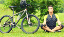 Man sitting next to his bicycle in a meditative pose with green hills, trees and grass