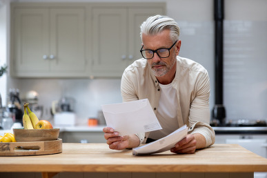 Man with gray hair and beard and mustache looking at a piece of mail in his kitchen