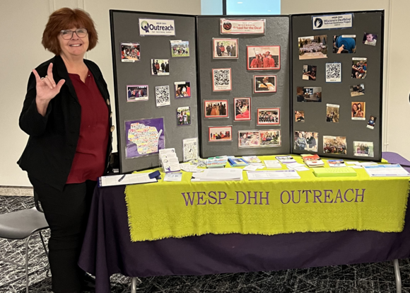 Maryann Barnett signs "I love you" with her right hand and poses with a poster board for the WESP-DHH Outreach.
