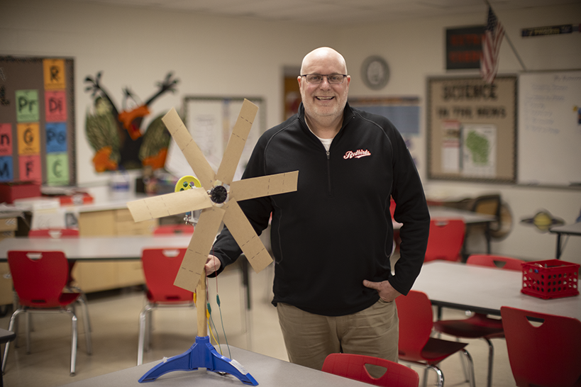 Mark Acherman smiles inside the classroom while showing a wind turbine school project made by his students.