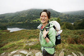 A smiling adult with a backpack standing on a hill front of a mountain