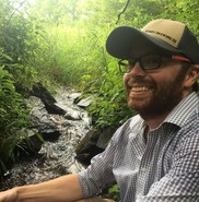 A photo of Ryan Jarvis in the woods with a stream flowing behind him.