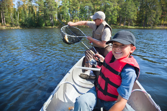 A kid smiles in a canoe while holding a fishing pole. An adult in the back of the canoe holds a net with a fish in it that the kid caught.