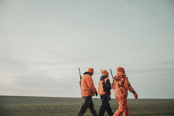 Three hunters wearing blaze orange walking across a field carrying rifles over their shoulders