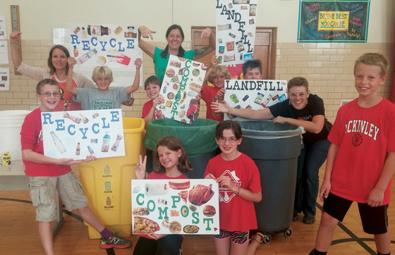 A group of elementary school students holding signs up saying "Recycle" and "Compost" in a school lunchroom.