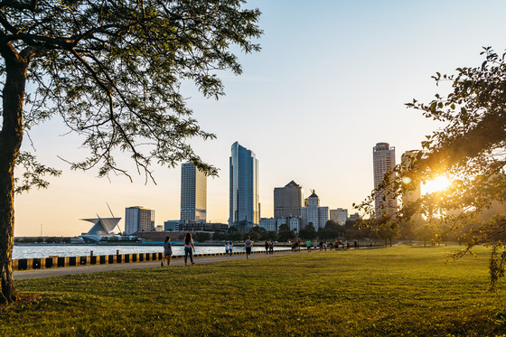 A skyline view of Milwaukee with people walking along footpath with building in the background while sun is setting. 