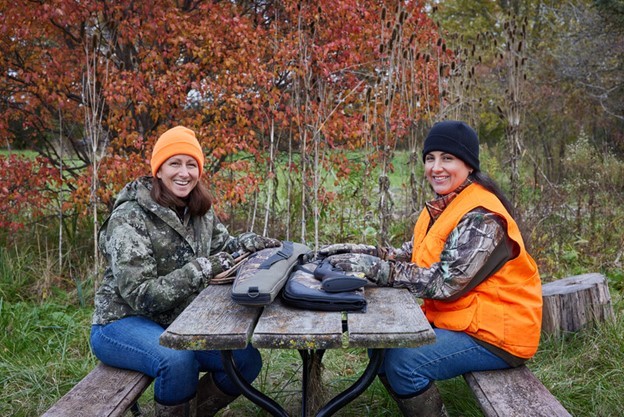 Two women sitting at a picnic table, dressed in hunting gear like camouflage vests and blaze orange hats.