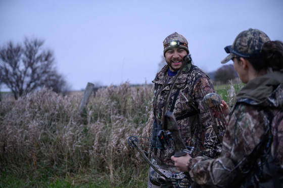 A male and female hunter wearing camouflage, holding bows in a field.