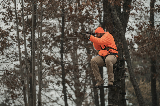 A male hunter sitting up in a tree stand, holding up his gun.