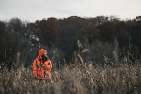 Woman hunter wearing blaze orange jacket and hat carrying a rifle over shoulder while standing in a field.