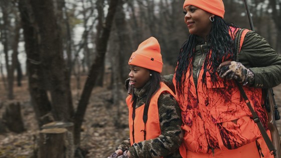 a younger woman wearing blaze orange and camo walks in the woods with an older woman wearing blaze orange and camo