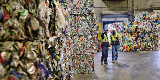 Two recycling plant workers walk through a warehouse lined with bales of recycled plastic.