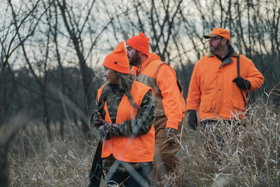 Three hunters walk through a field of tall grass safely shouldering their firearms and wearing blaze orange.