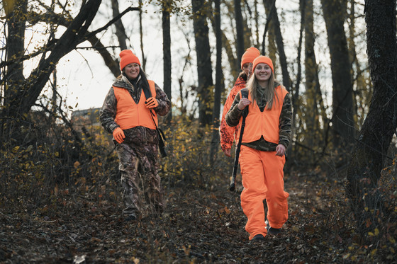 group of three women wearing blaze orange carrying rifles walking through the woods