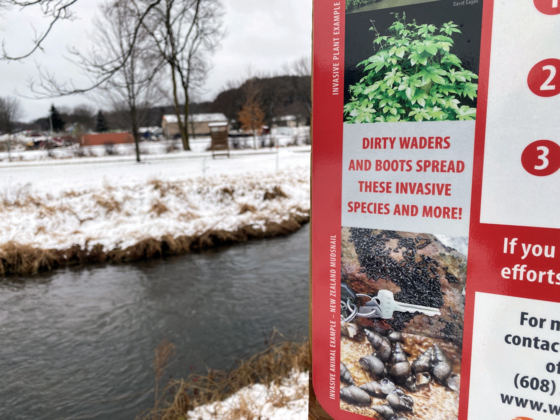 A sign with tips on how to avoid aquatic invasive species in front of a stream with snow on the ground.