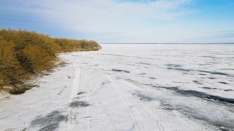 Snowmobile tracks cross the frozen water. Snow covers the lake surface, and a bright blue sky above.