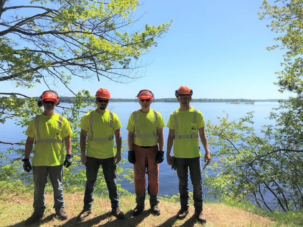 A group of four parks workers in yellow shirts and orange hats, standing by trees and a body of water in the sunshine.