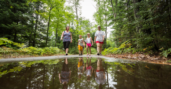 two adults and three children walking through the woods on a paved trail. There is a puddle on the ground in front of them.