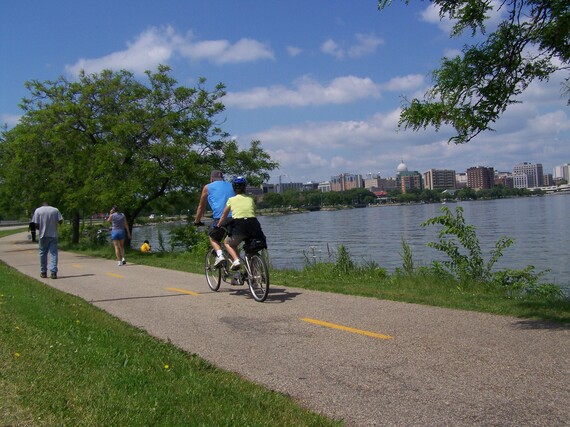Tandem bike riders and walkers on paved path with lake and Madison skyline in background