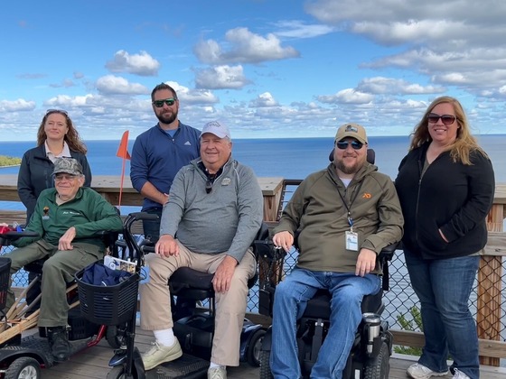 Six people posing for a photo at the top of Eagle Tower at Peninsula State Park with Lake Michigan and fluffy white clouds in the background.