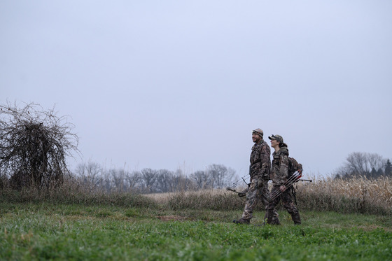 a man and woman wearing camo walk across a field carrying crossbows