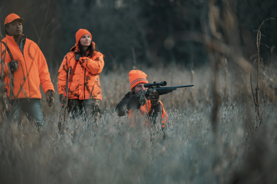 Three hunters, all in blaze orange, hunting in a dried cornfield. The hunter in the front of the group points her rifle at a target in the distance. 