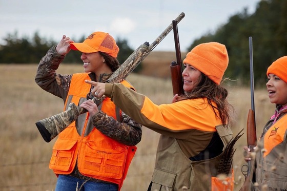 Three women, all wearing Blaze Orange, hunting in a cornfield. One woman points at a target outside of the frame. 