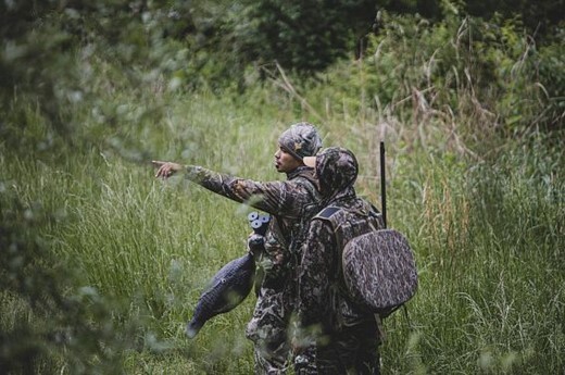 two men wearing camo trek through the woods carrying hunting gear