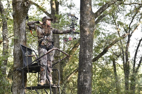 A young woman wearing camouflage draws her bow and takes aim while hunting from a treestand. 