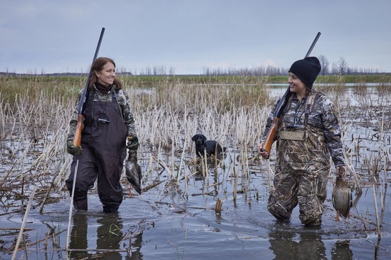 Two women, both standing in knee-deep water, smile at each other after a successful hunt. A black dog stands behind them. 