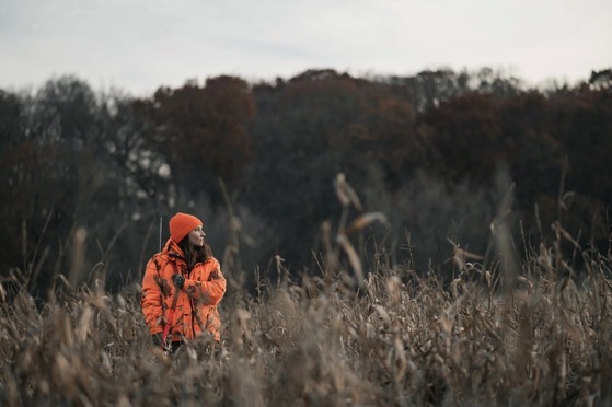 A young woman, wearing blaze orange, stands in a dried cornfield while hunting for deer. 