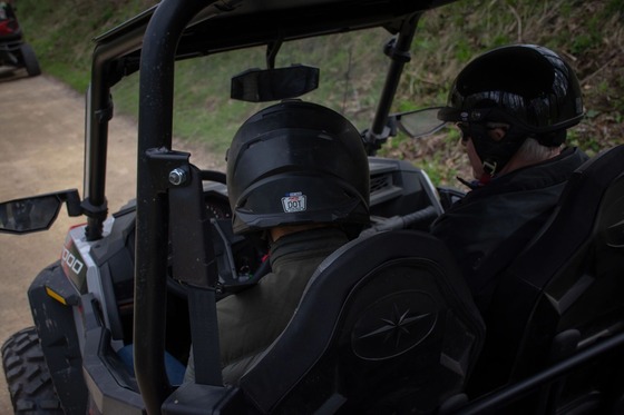 Two people, both wearing black helmets, dark jackets and wearing their seatbelts, riding a UTV to get to their hunting spot. 