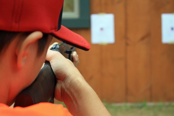 Viewed from behind, a teenage boy, wearing a blaze orange shirt and red hat, aims his rifle at a target at a hunter education event. 