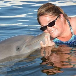 A woman standing in the ocean smiles as she greets a dolphin.