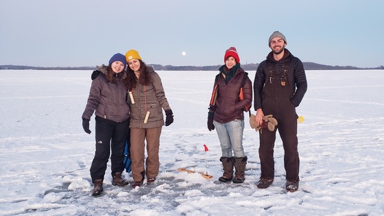 Four individuals pose for a picture around a tip up while ice fishing. 