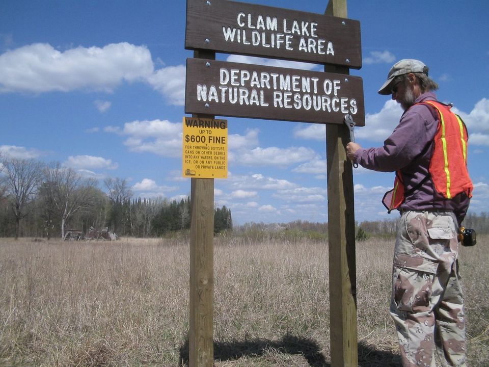 A DNR employee, wearing an orange vest and holding tools, performs maintenance on a trail sign at Clam Lake Wildlife Area. 