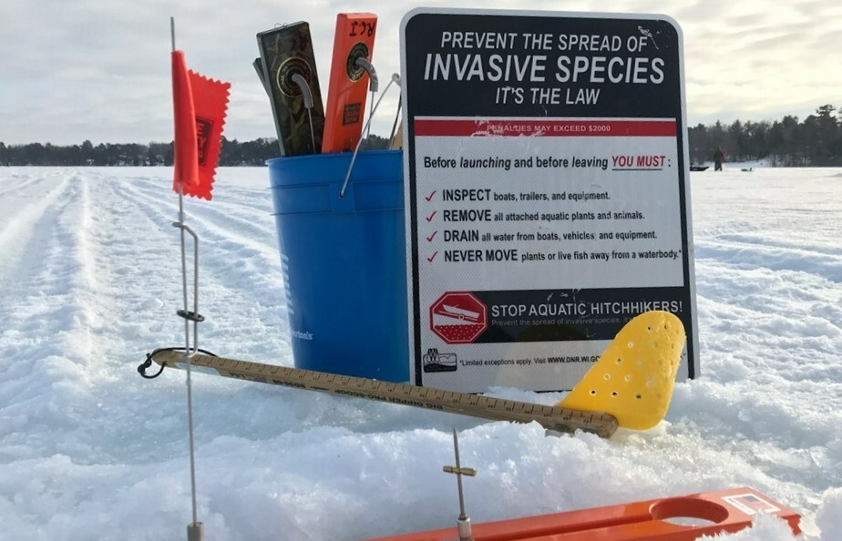 A sign that says "prevent the spread of invasive specie - it's the law" sits next to a bucket of ice fishing equipment on a frozen lake. 